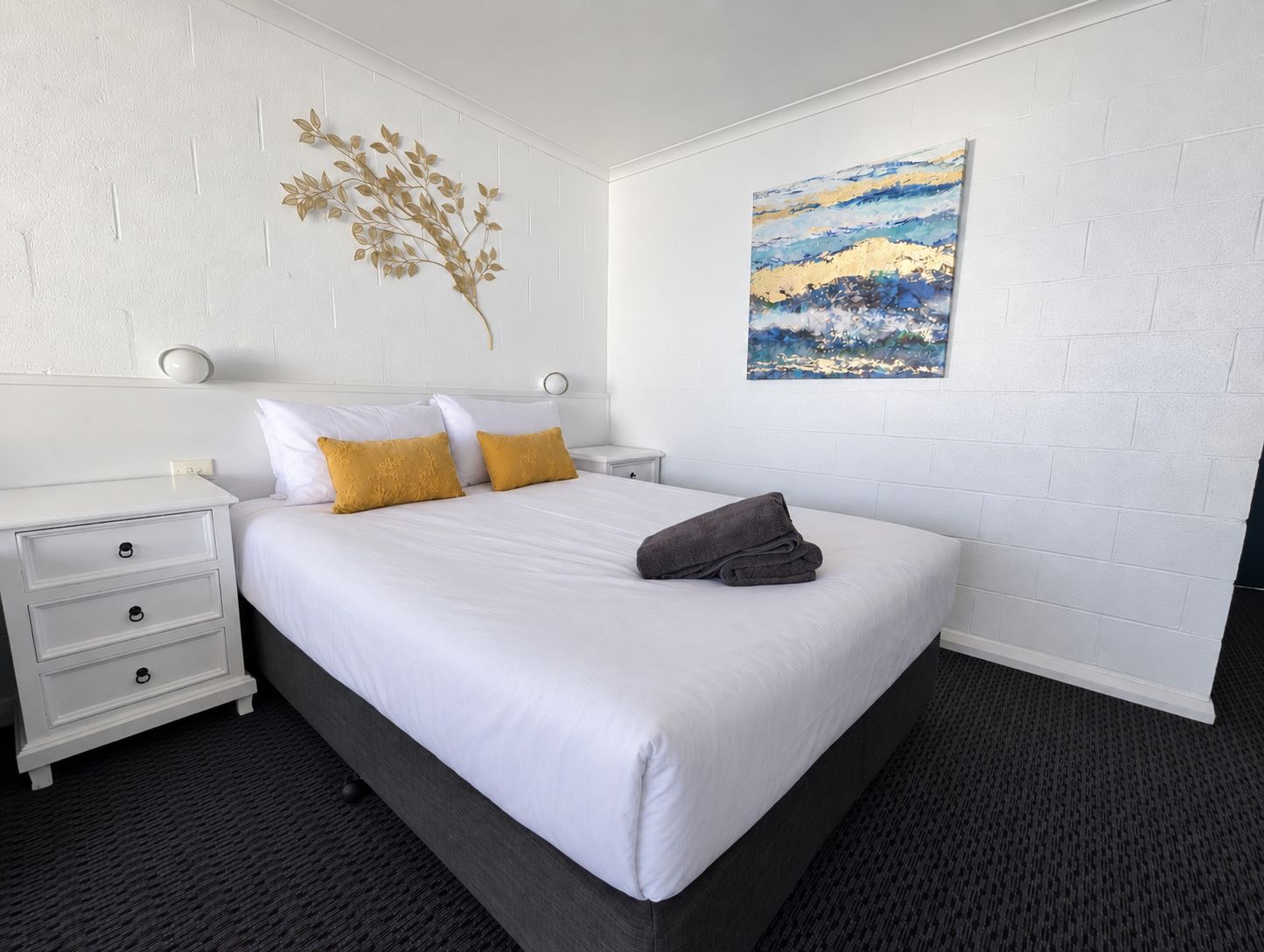Une chambre d’hôtel avec un lit, des tables de chevet blanches, une plante fixée au mur et un tableau.