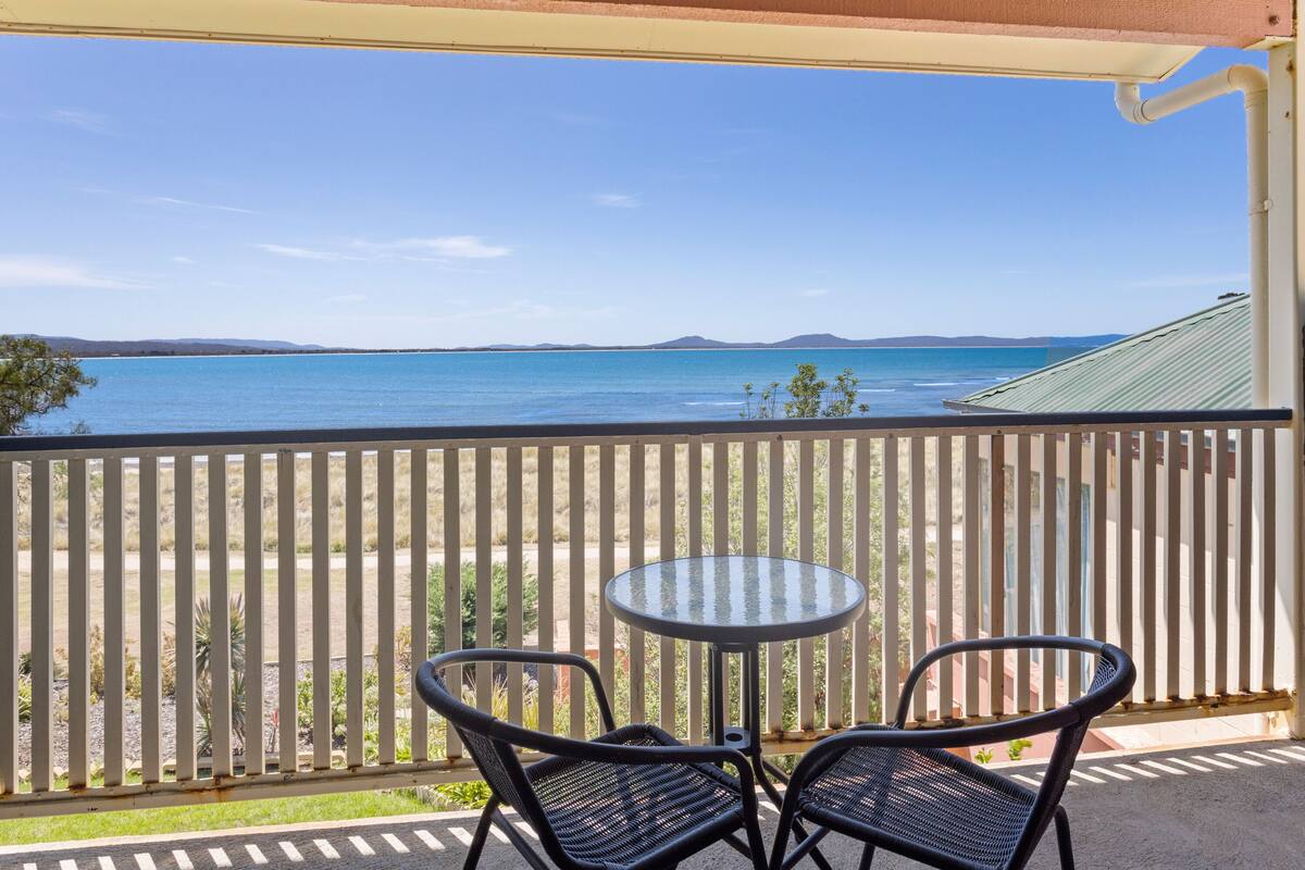 A balcony with a table and two chairs, overlooking a body of water and distant land.
