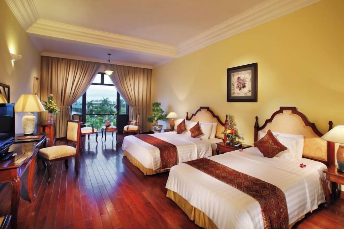 A hotel room with two beds, a desk with a lamp, a television, and a view of greenery outside.