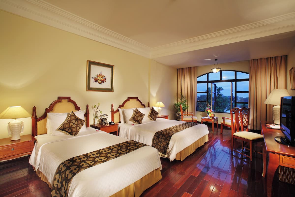 A hotel room with two beds, wooden headboards, a desk with a television, and a large window with curtains.