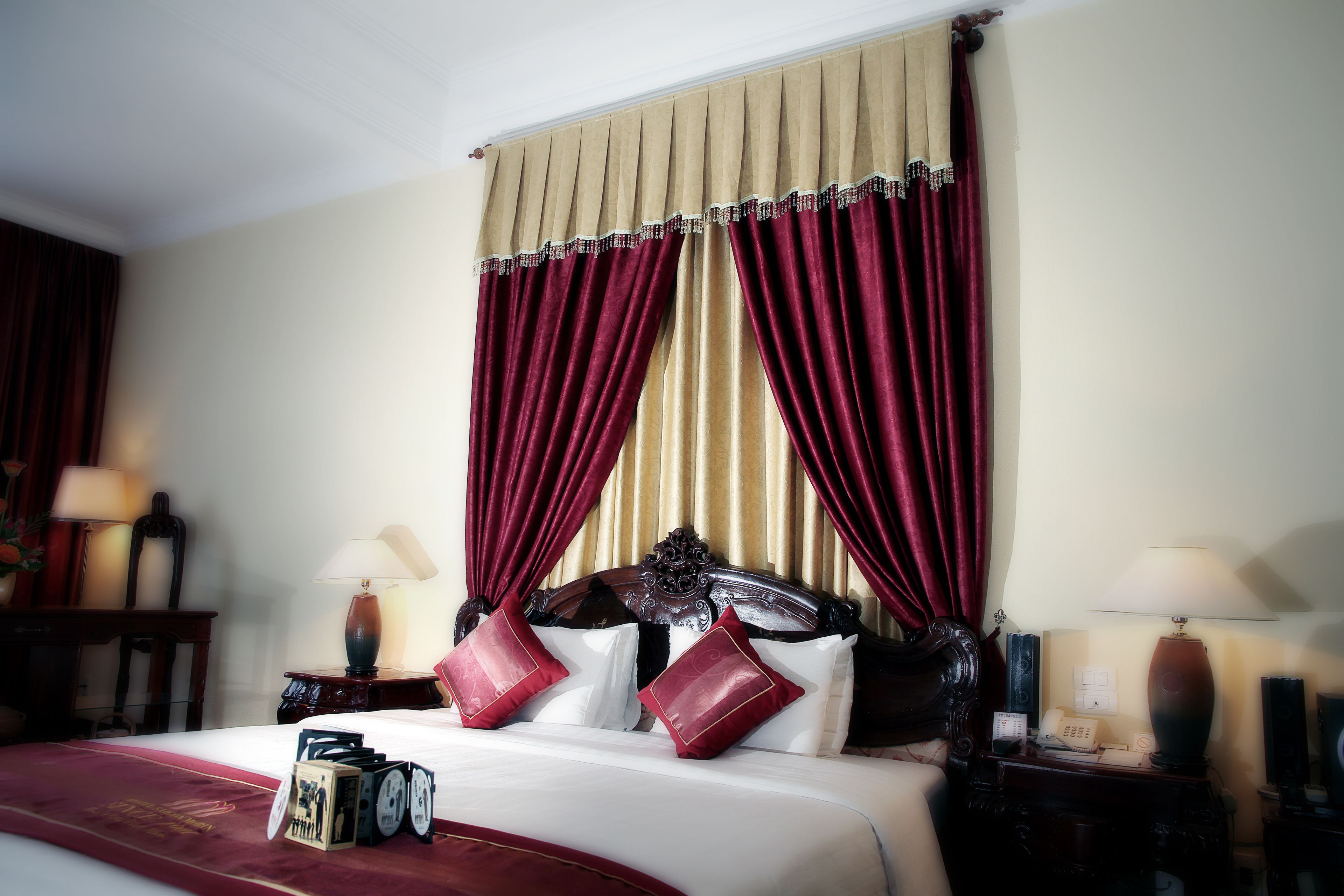 Quarto de hotel com cama grande, cortinas vermelhas e bege, cabeceira de madeira e mesa de cabeceira com luminária e telefone.