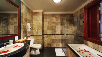 A bathroom with a marble wall, a bathtub with rose petals, a glass shower enclosure, and a sink with a mirror.