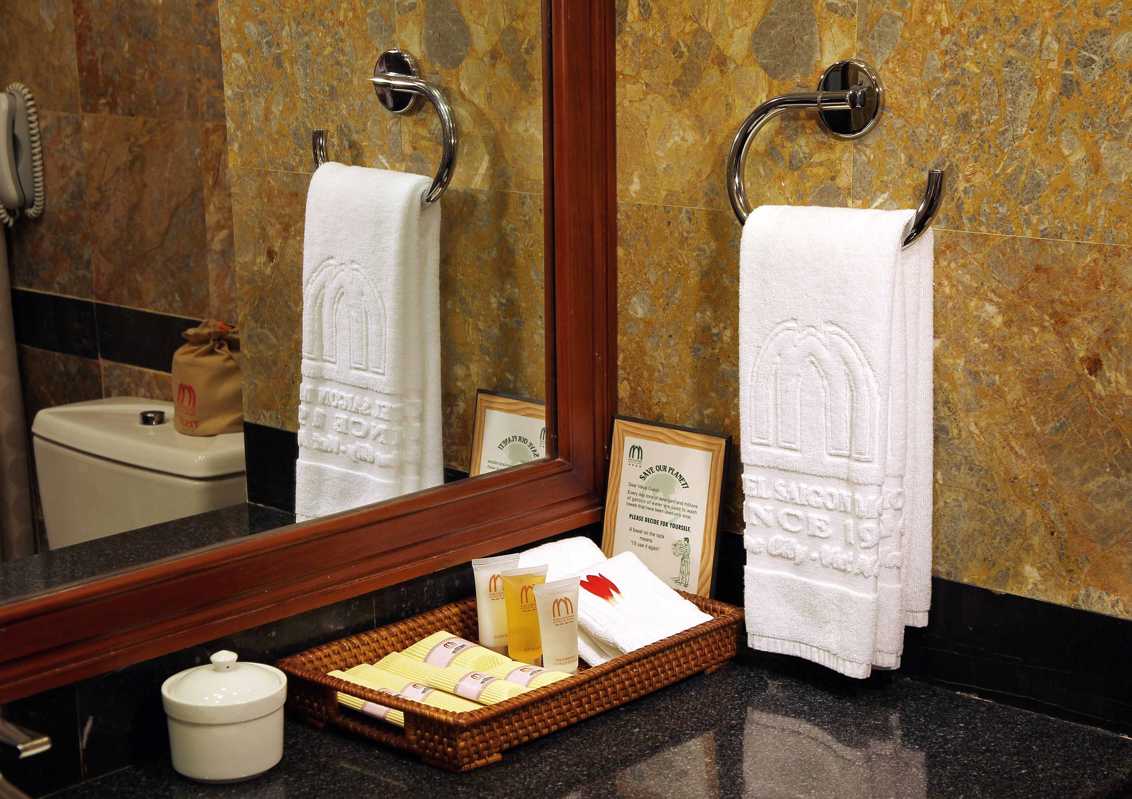A bathroom with two white towels hanging on towel bars, a mirror reflecting the towels and a framed welcome sign, a wicker basket with toiletries, and a white ceramic container.