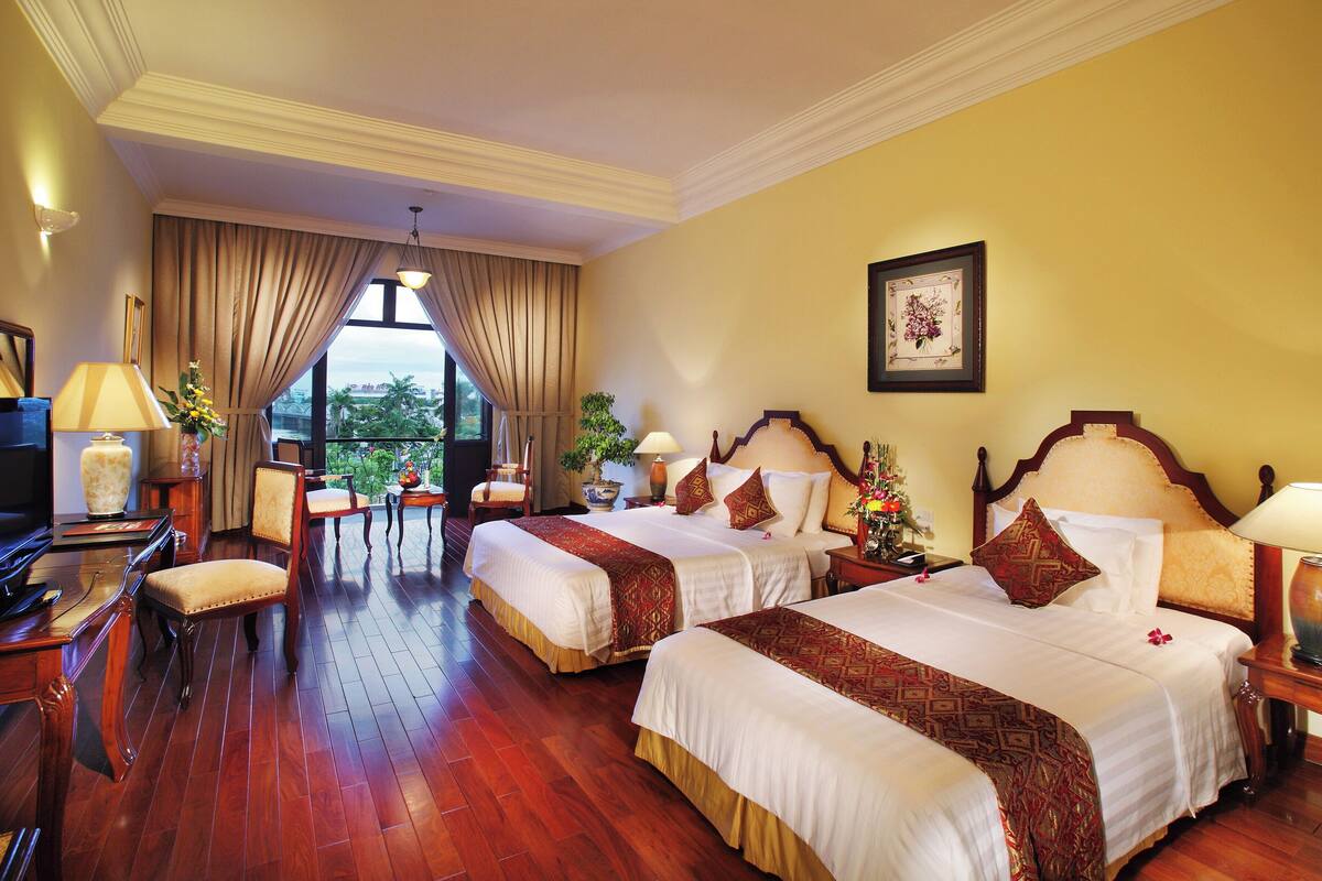 A hotel room with two beds, a desk with a TV, and a view of greenery outside.