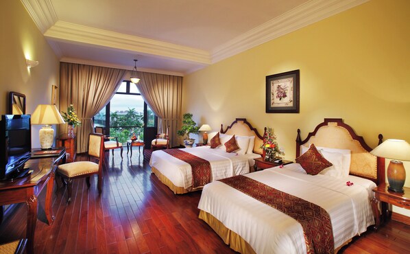 A hotel room with two beds, a desk with a TV, and a view of greenery outside.