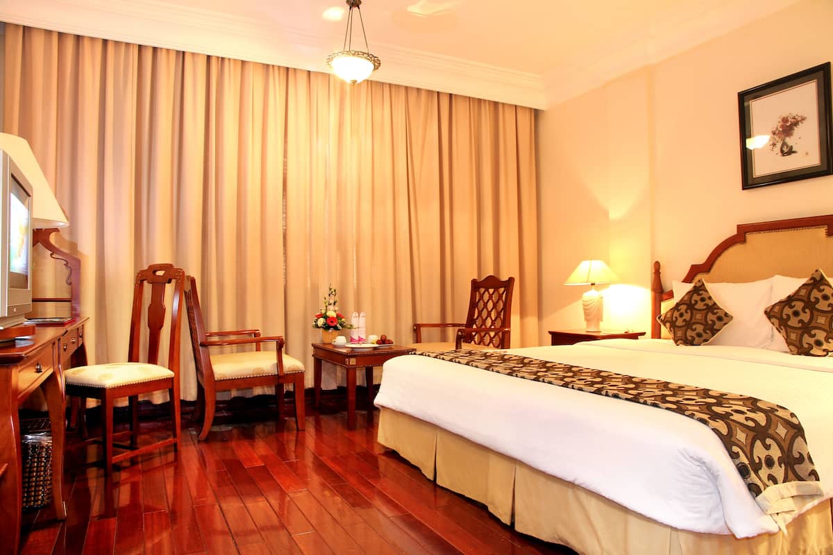 A hotel room with a large bed, two wooden chairs, a desk with a lamp, and a large window with curtains.