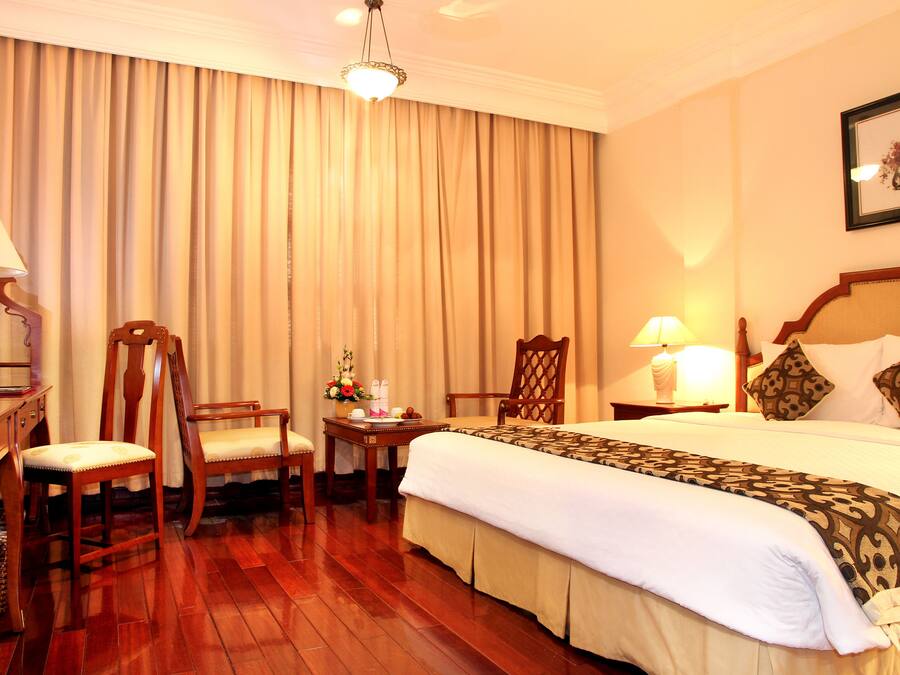 A hotel room with a large bed, two wooden chairs, a desk with a lamp, and a large window with curtains.