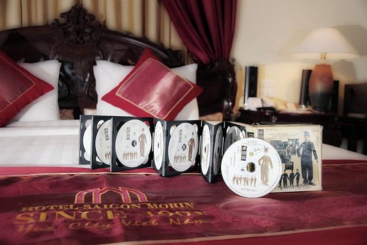 A neatly made bed with red and white pillows, a red bedspread with the hotel's name, and a set of CDs or DVDs on the bed.