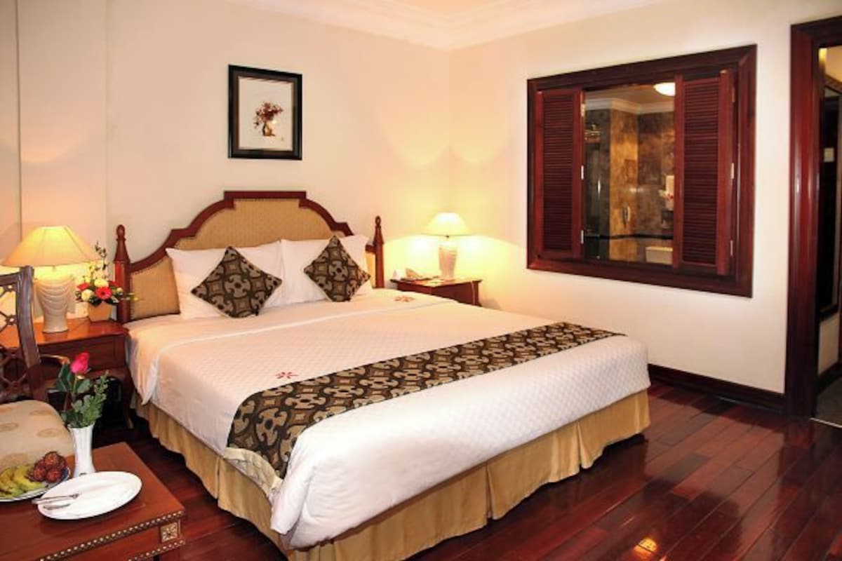 A neatly arranged hotel room with a large bed, a bedside table with a lamp, a framed picture on the wall, and a window with wooden shutters.