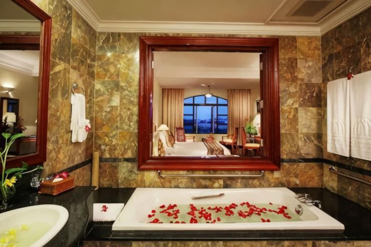 A bathroom with a large bathtub, a mirror, and a view of a bedroom with a bed and window.