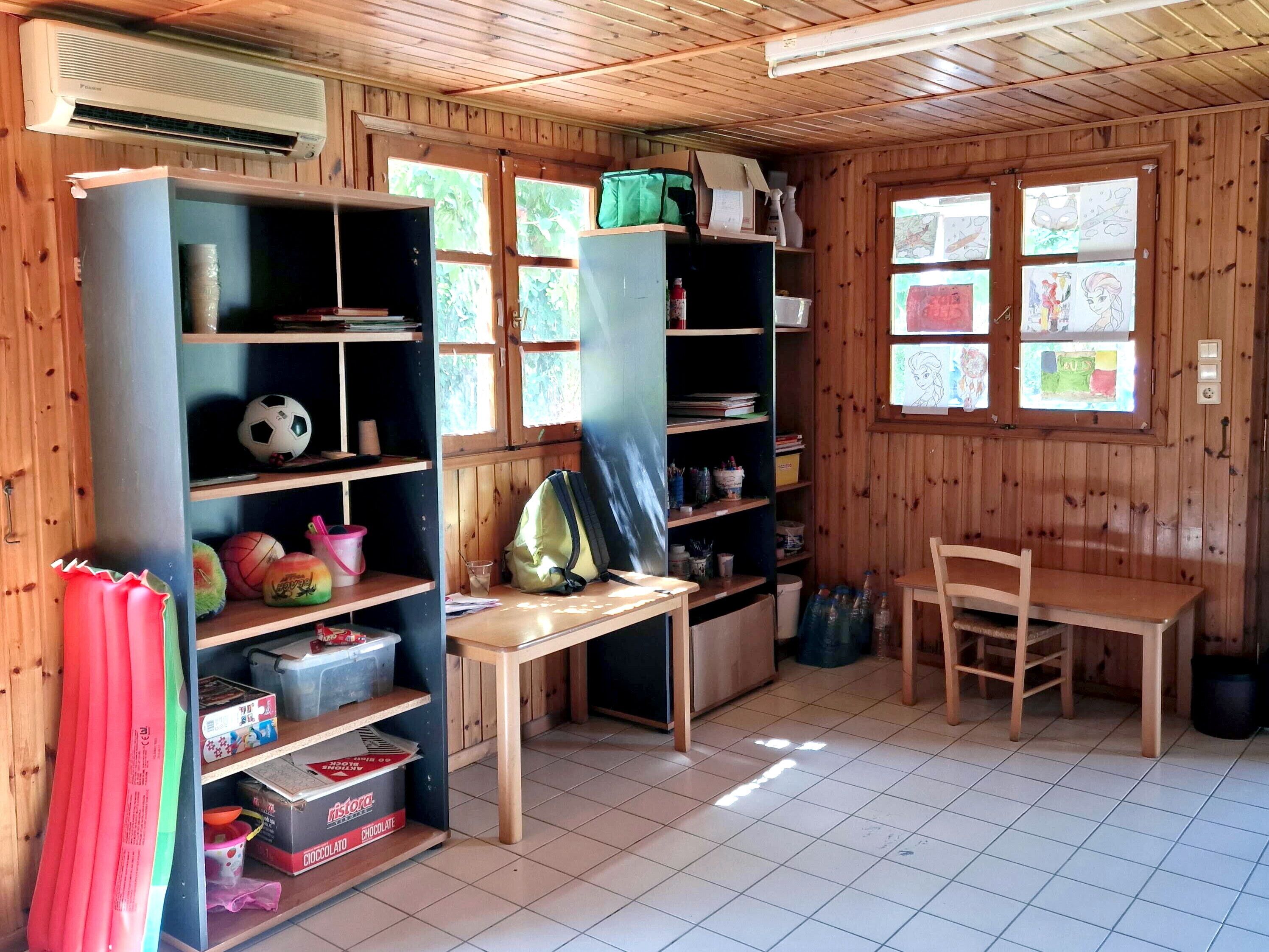 Una habitación con paredes y techo de madera, suelo de baldosas y estantes con diversos objetos, incluyendo balones deportivos, libros y una bolsa verde.
