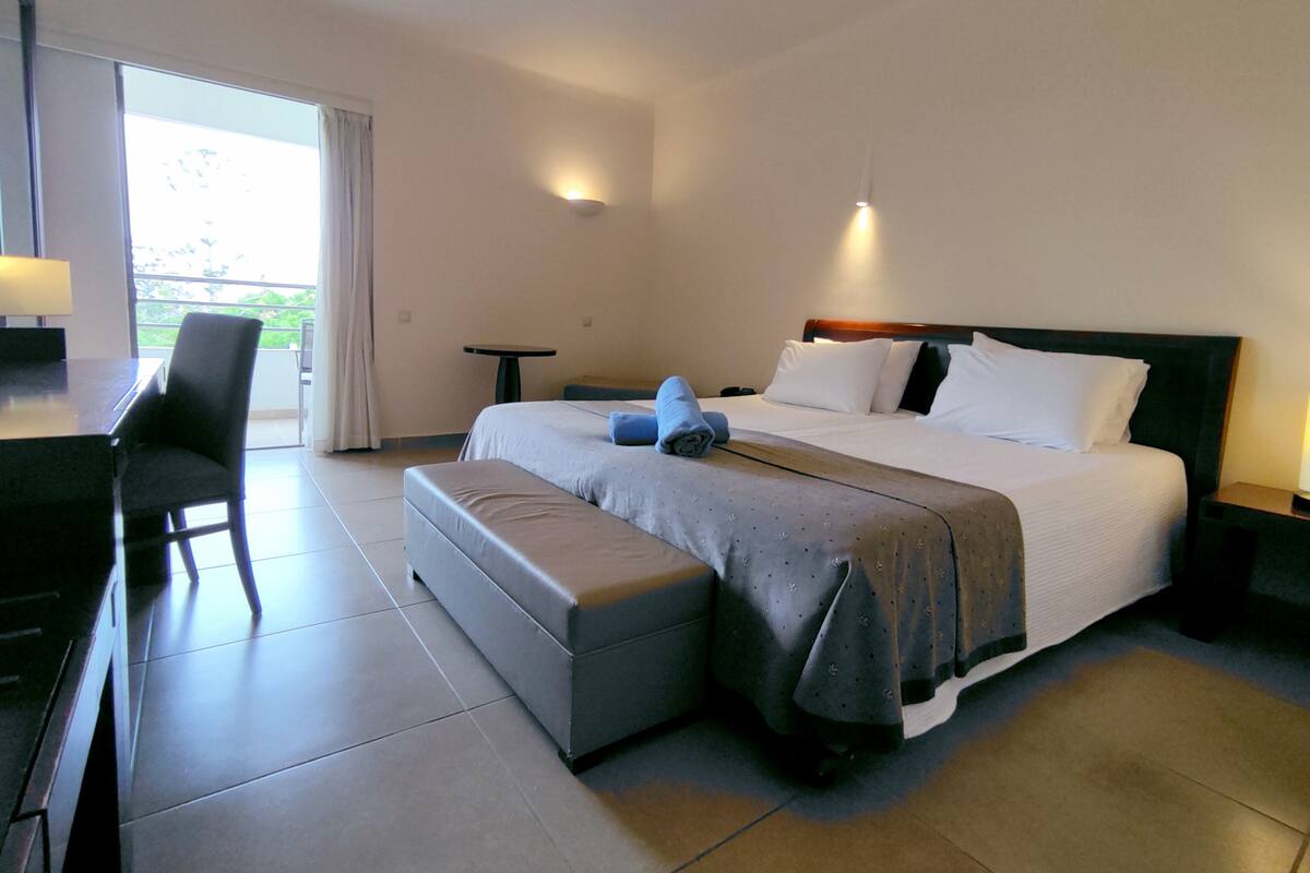 A hotel room with a bed, a desk, a chair, a small table, and a balcony view.