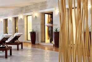 A hotel lobby with wooden lounge chairs, large potted plants, and a reception area with a person holding a white object. - Apollonia Beach Resort & Spa (Malevizi)