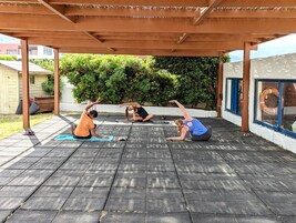 Un espacio al aire libre cubierto con un pérgola de madera, colchonetas de ejercicio y follaje al fondo.