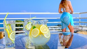 A table with a pitcher and glasses of lemonade, a person in a blue swimsuit, and a view of the ocean. - Apollonia Beach Resort & Spa (Malevizi)