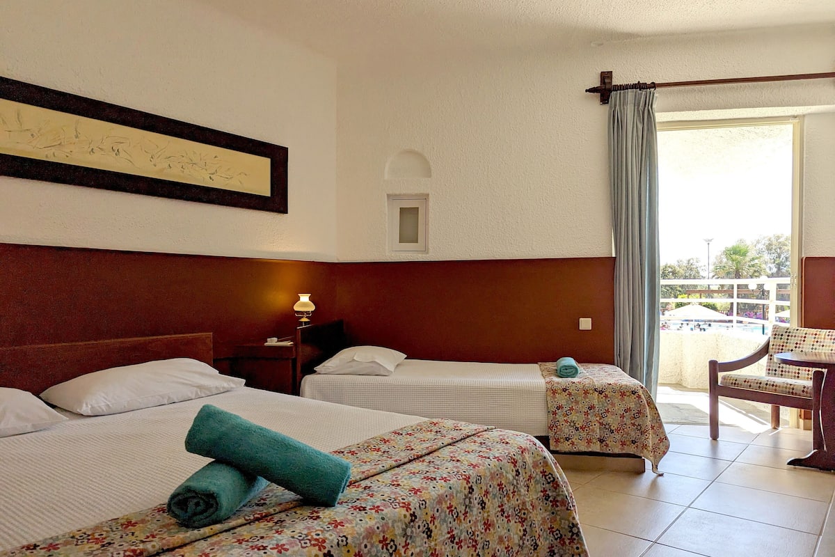 A hotel room with two beds, a small table with a lamp, a chair, and a balcony with a view of the pool.