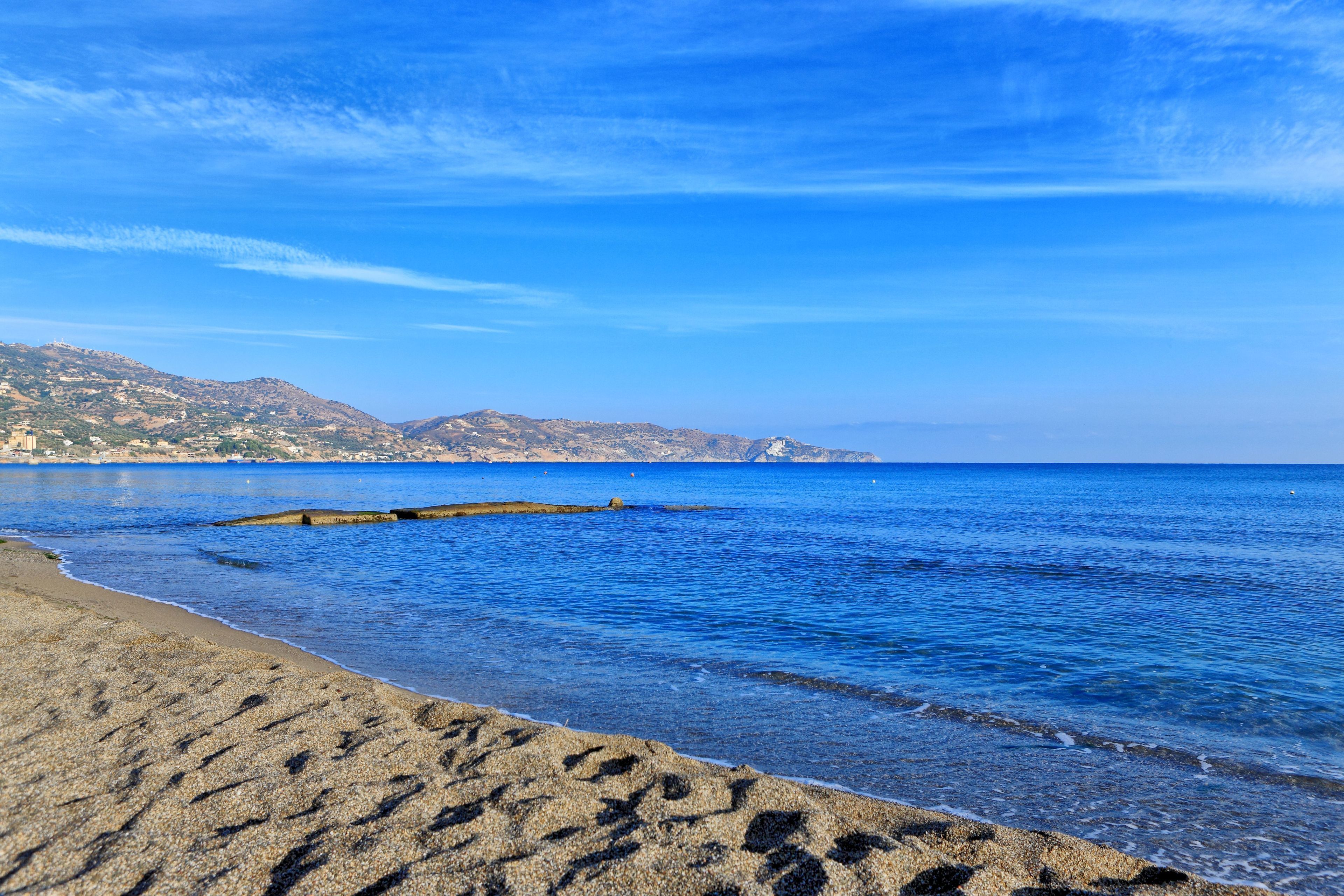 A beach with clear blue water, a rocky shoreline, and distant hills.