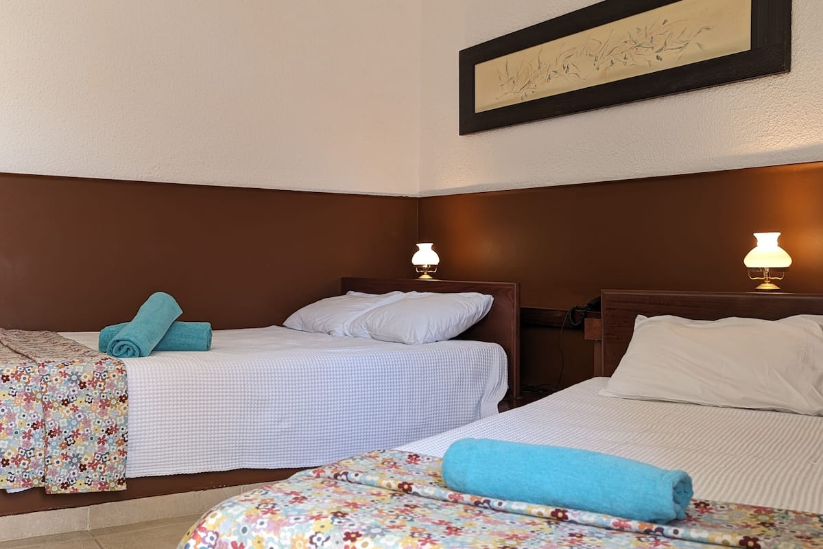 A hotel room with two beds, each with a white bedspread and a blue towel. There are two wall-mounted lamps and a framed artwork above the beds.