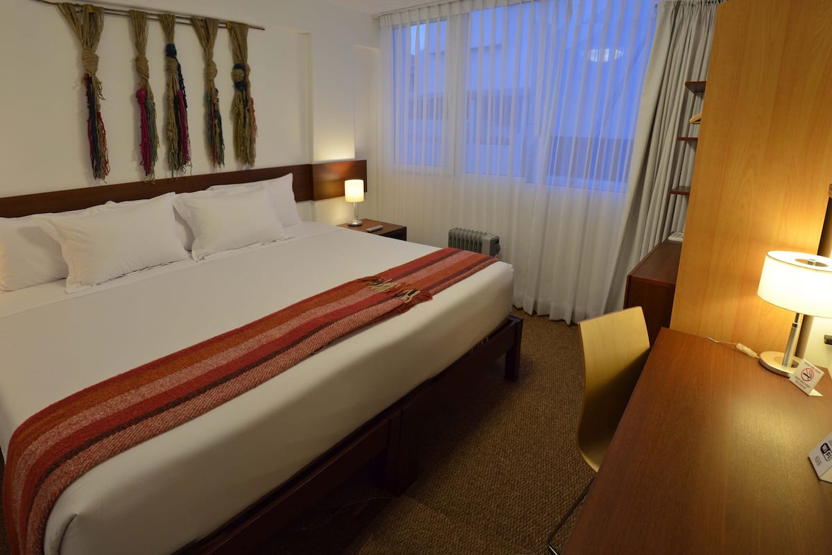 A hotel room with a large bed, two bedside tables with lamps, a wooden headboard, a wall-mounted telephone, and a window with sheer curtains.