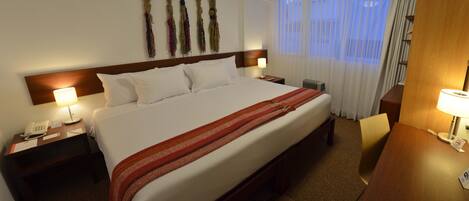 A hotel room with a large bed, two bedside tables with lamps, a wooden headboard, a wall-mounted telephone, and a window with sheer curtains.