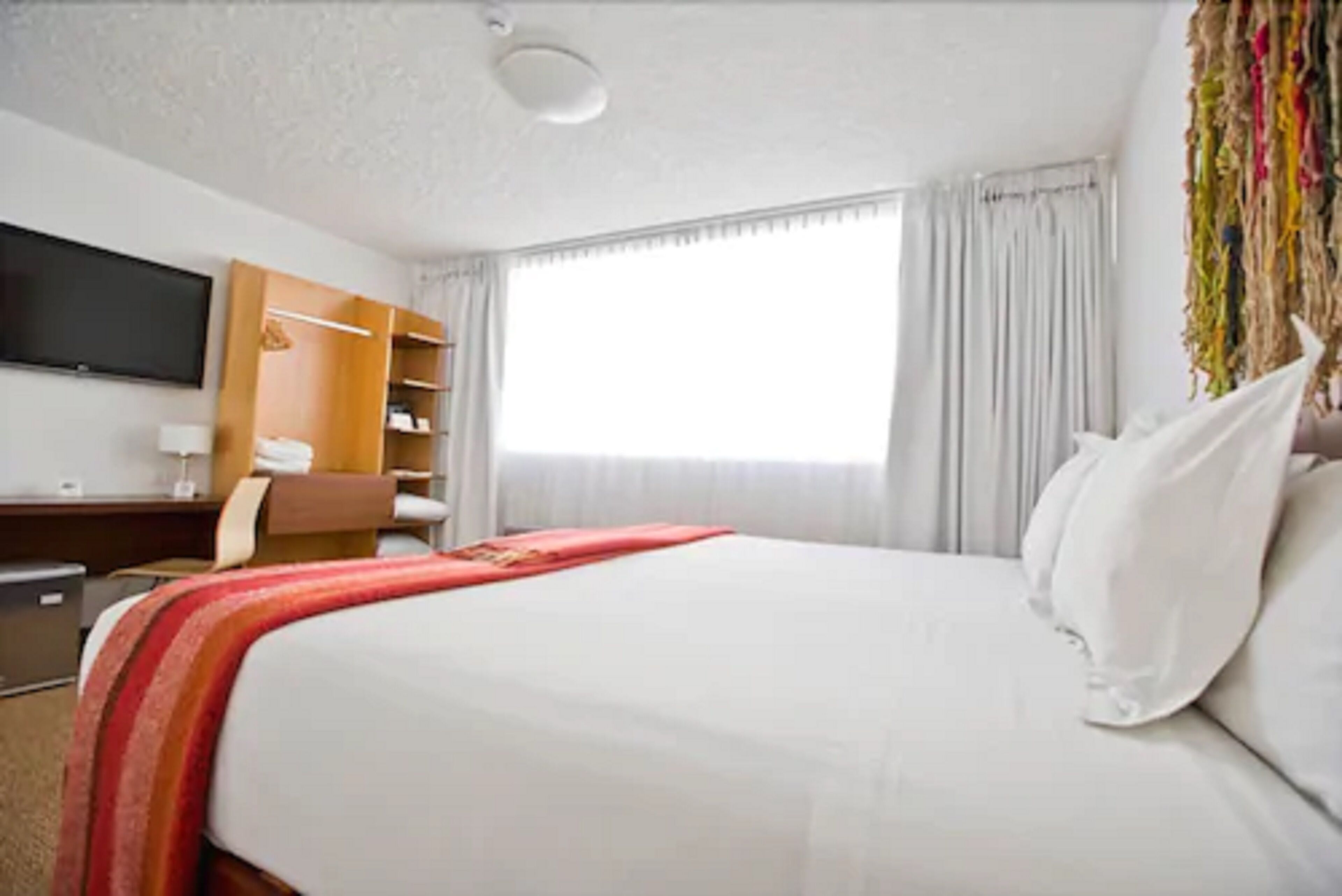 A hotel room with a bed, a TV mounted on the wall, a desk with a chair, and a wardrobe.