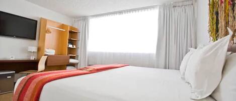 A hotel room with a bed, a TV mounted on the wall, a desk with a chair, and a wardrobe.