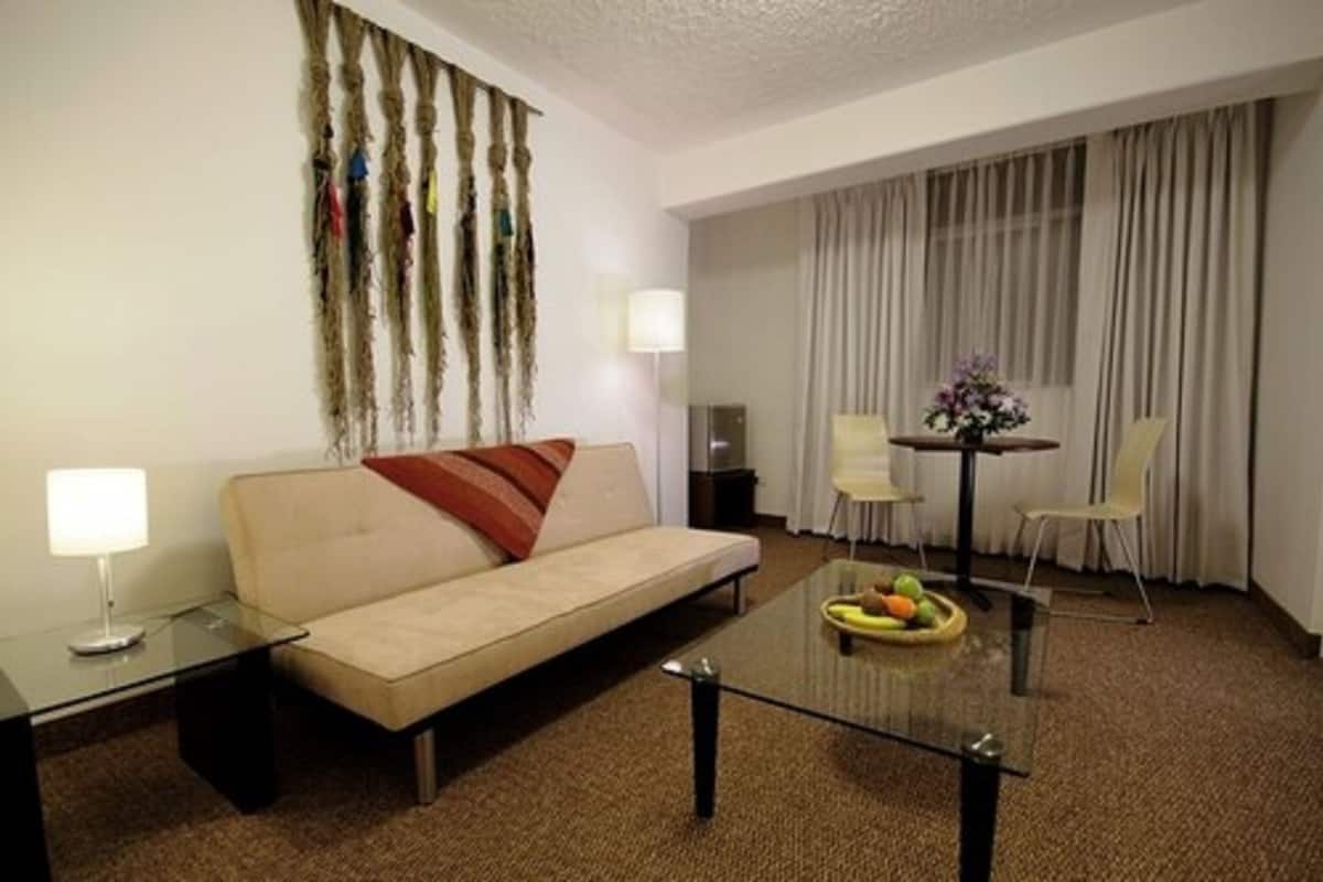 A hotel room with a sofa, a glass coffee table with fruit, a small table with a lamp, and a chair.