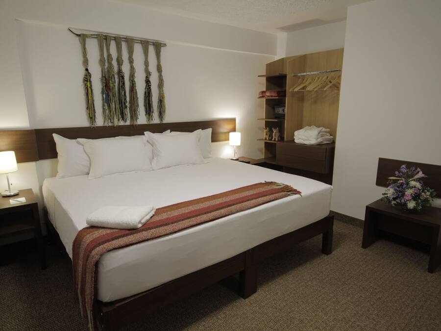 A hotel room with a large bed, two bedside tables with lamps, a wooden headboard, a wardrobe, and a small nightstand with a vase of flowers.