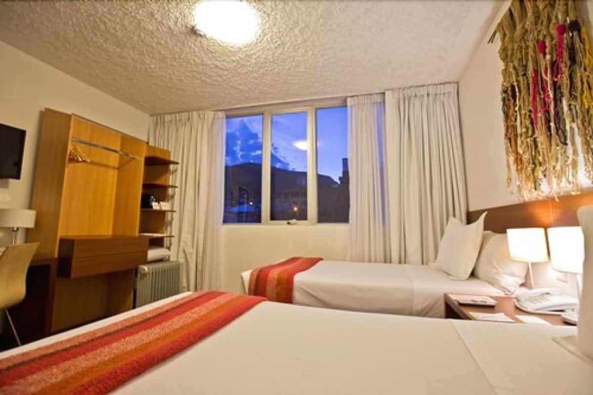 Une chambre d’hôtel avec deux lits, une armoire en bois, un bureau avec une lampe et une vue sur les montagnes par la fenêtre.