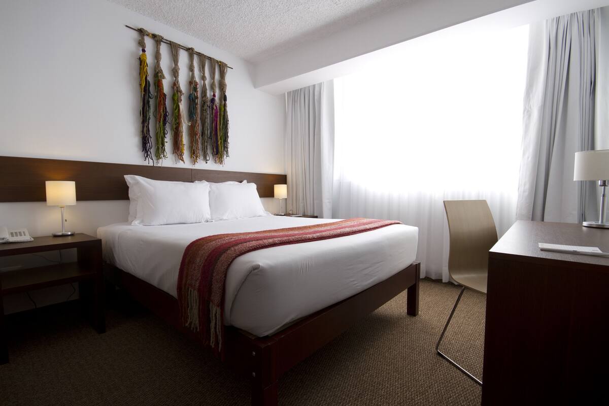A hotel room with a large bed, bedside tables, a desk, and a window with curtains.