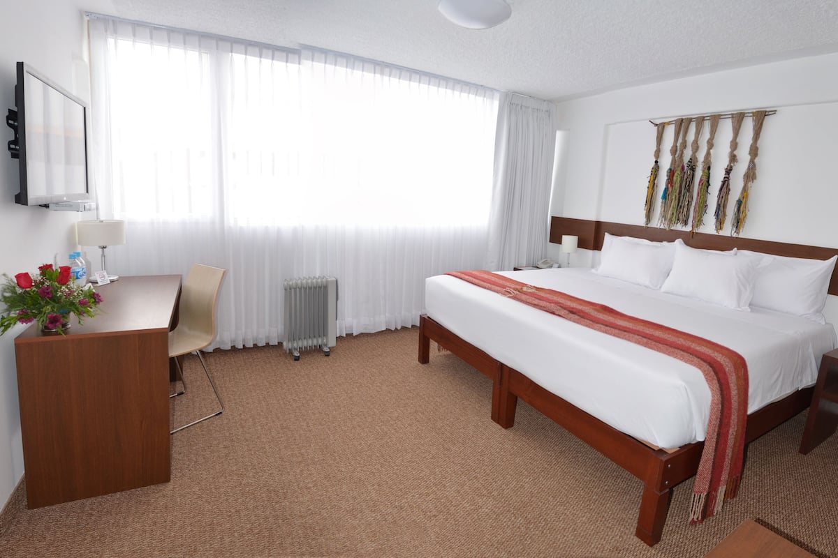 A hotel room with a large bed, a desk, a chair, a radiator, and a wall-mounted TV.