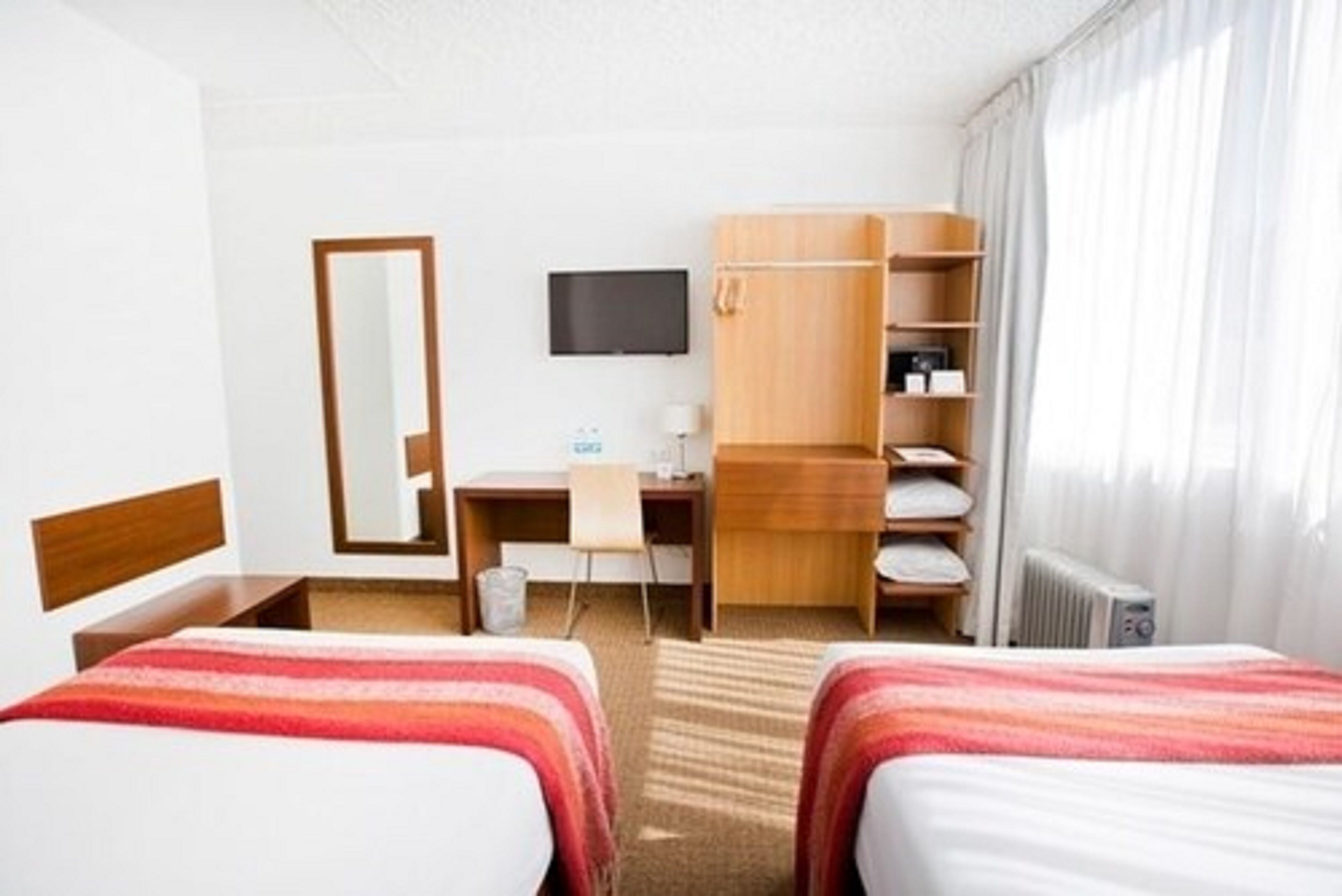 A hotel room with two beds, a desk, a chair, a TV, a wardrobe, and a radiator.