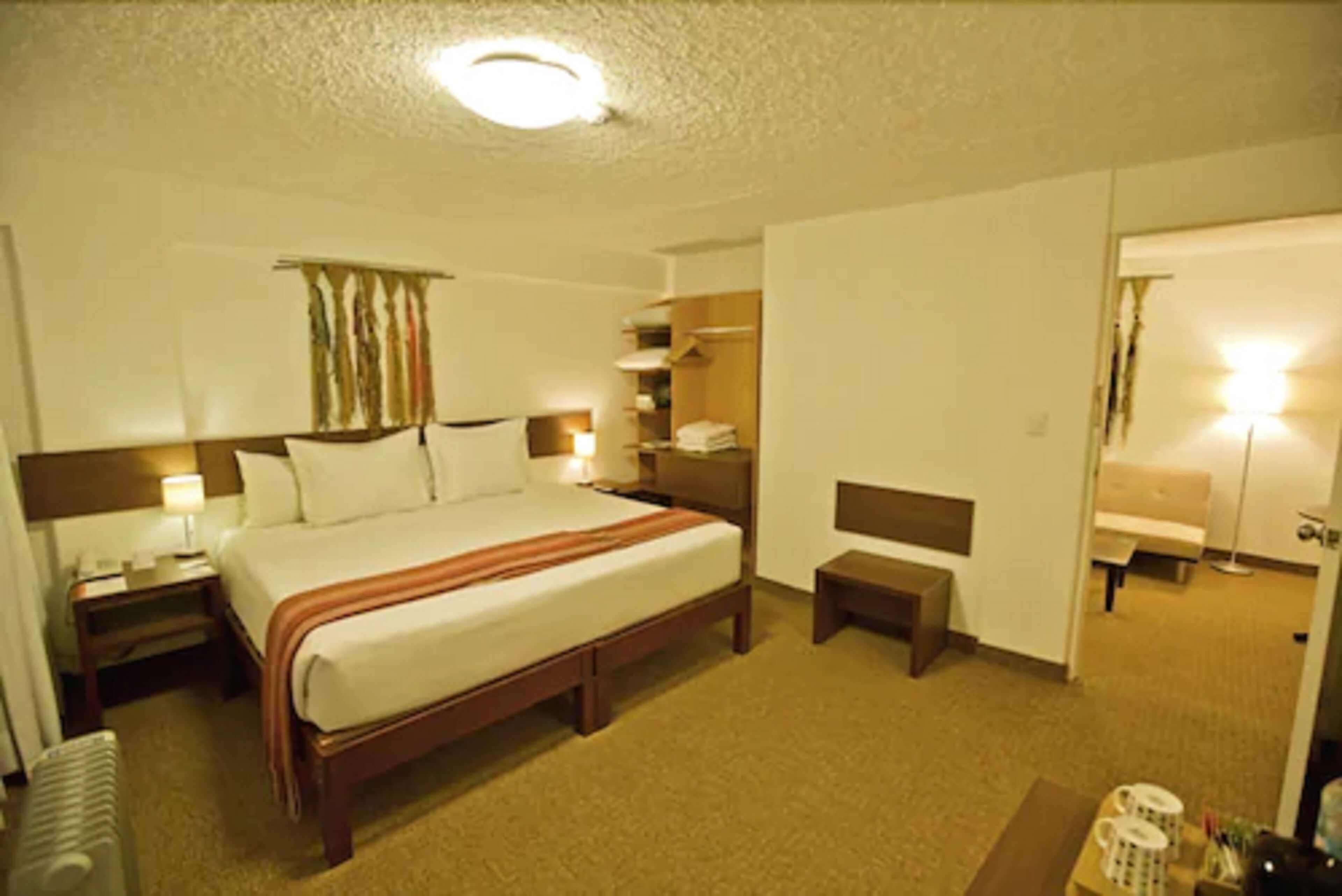 A hotel room with a large bed, bedside tables, a nightstand, a bench, a wardrobe, and a mirror.