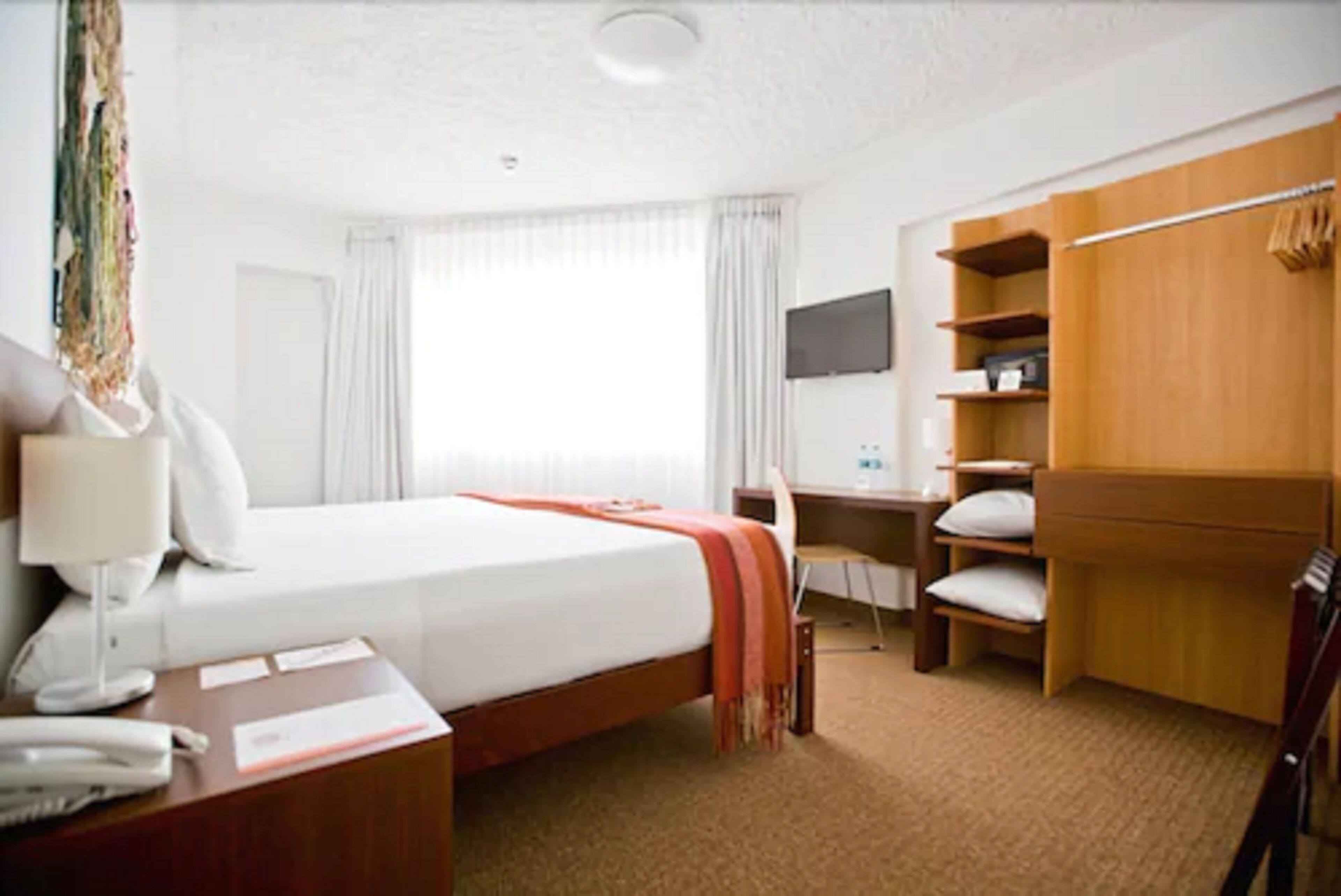 A hotel room with a bed, bedside table, desk, chair, wardrobe, and a television on the wall.