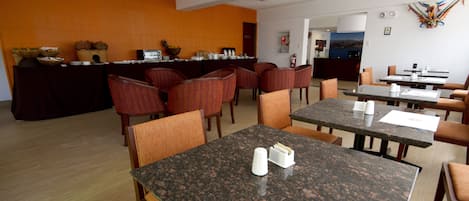A hotel dining area with multiple tables and chairs, a reception desk, and a kitchen area.