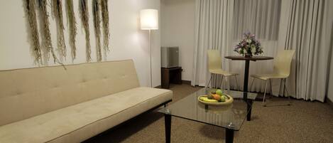 Un salon comprenant un canapé, une table basse en verre, deux fauteuils, une lampe et une petite table sur laquelle se trouvent des fruits.