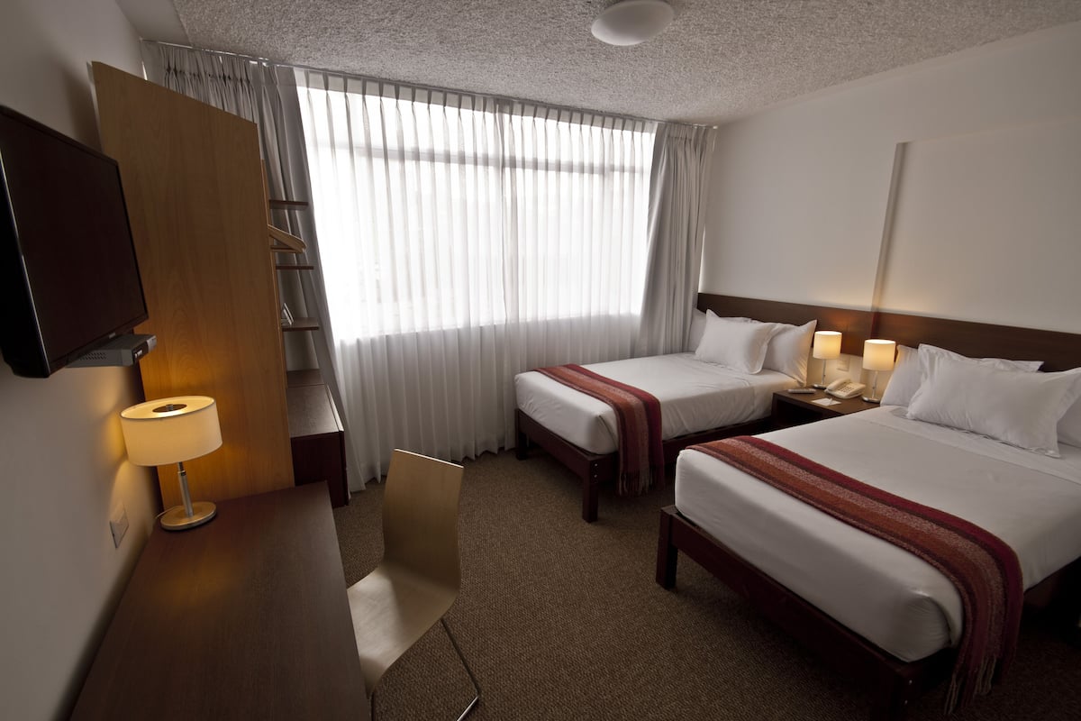 A hotel room with two beds, a desk with a lamp, a TV, and a window with curtains.
