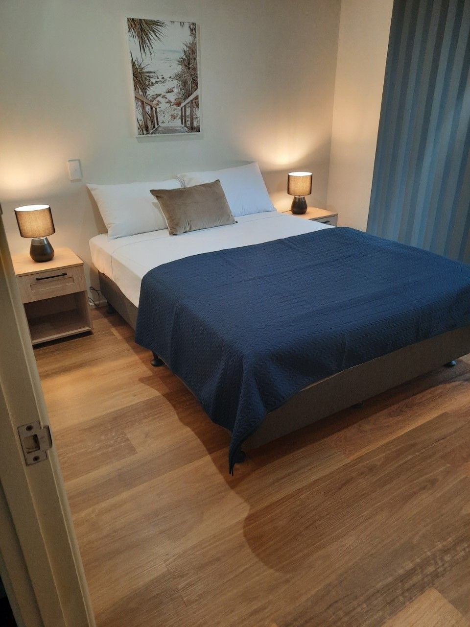 A neatly made bed with a blue bedspread, two bedside lamps, a nightstand, and a wall-mounted picture.