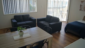 Um quarto com uma mesa de jantar de madeira, dois sofás escuros, uma cama com cobertura azul e uma janela com persianas.