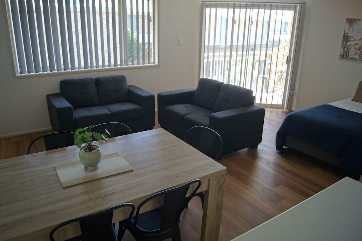 Une pièce comprenant une table à manger en bois, deux canapés sombres, un lit recouvert d’une couverture bleue et une fenêtre avec des stores.