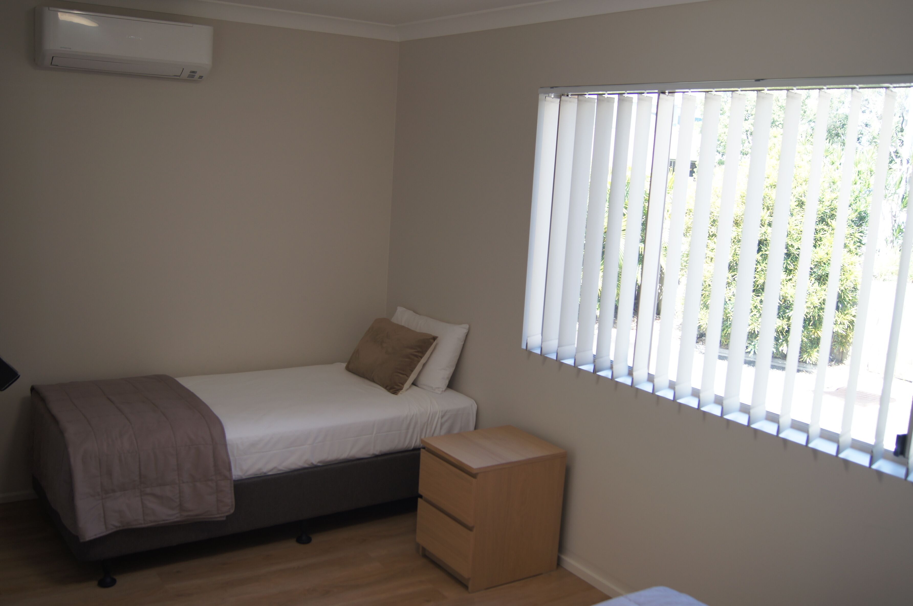 A small, single-bed room with a wooden floor, a bedside table, a window with blinds, and an air conditioning unit on the wall.