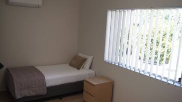 A small, single-bed room with a wooden floor, a bedside table, a window with blinds, and an air conditioning unit on the wall.