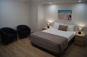 A bedroom with a bed, two chairs, a nightstand with a lamp, and a painting on the wall.