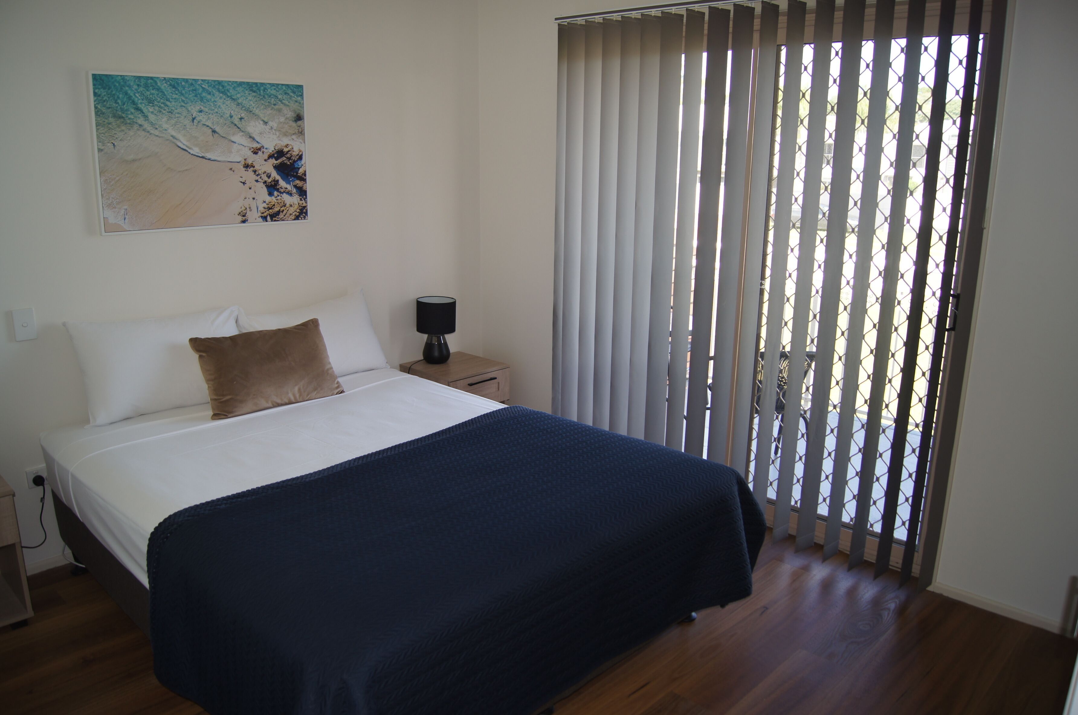 A bedroom with a bed, a nightstand, a lamp, a painting, and a window with blinds.