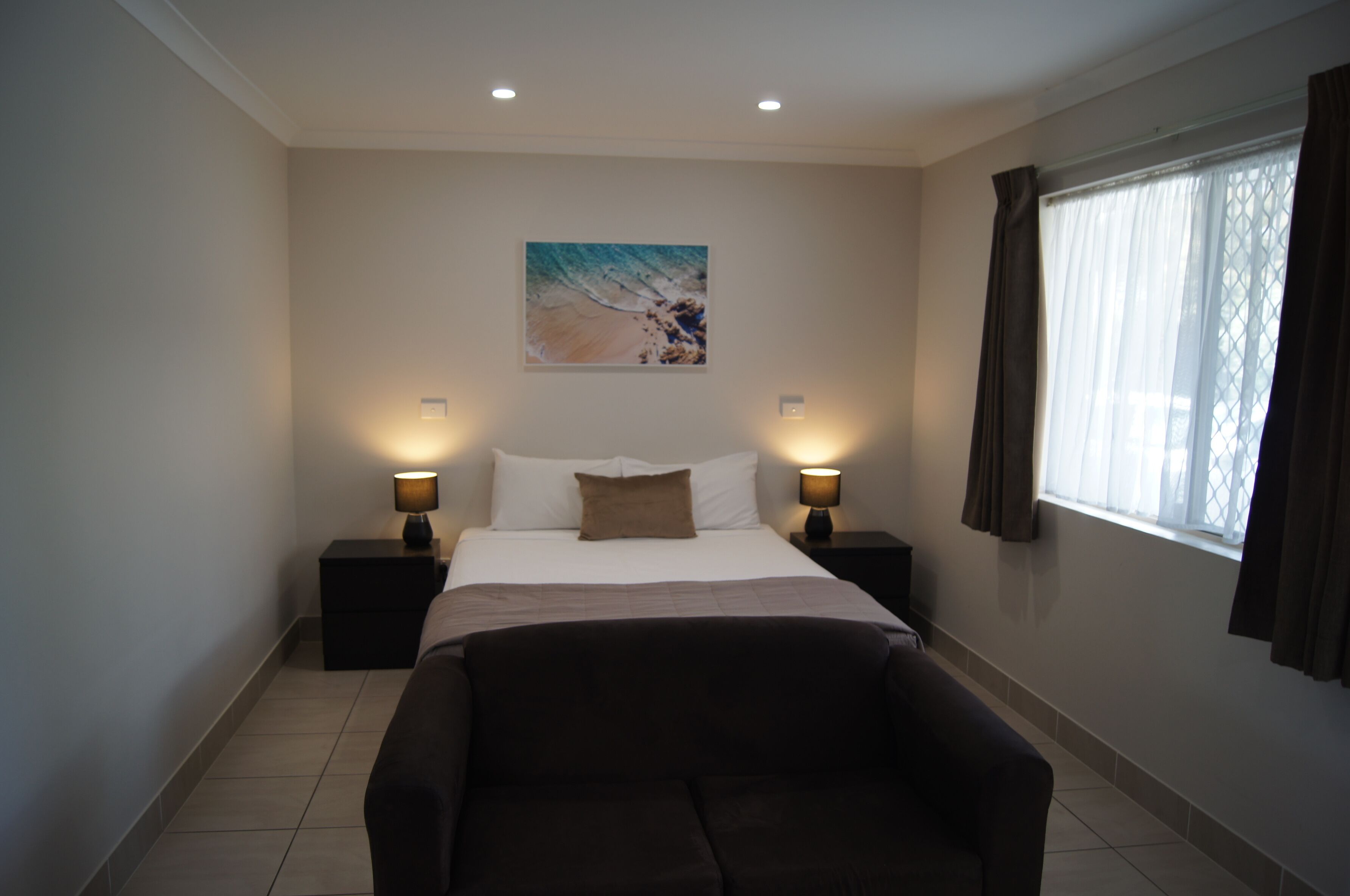Habitación de hotel con una cama, dos mesitas de noche con lámparas, un cuadro grande en la pared, una ventana con cortinas y un sofá.