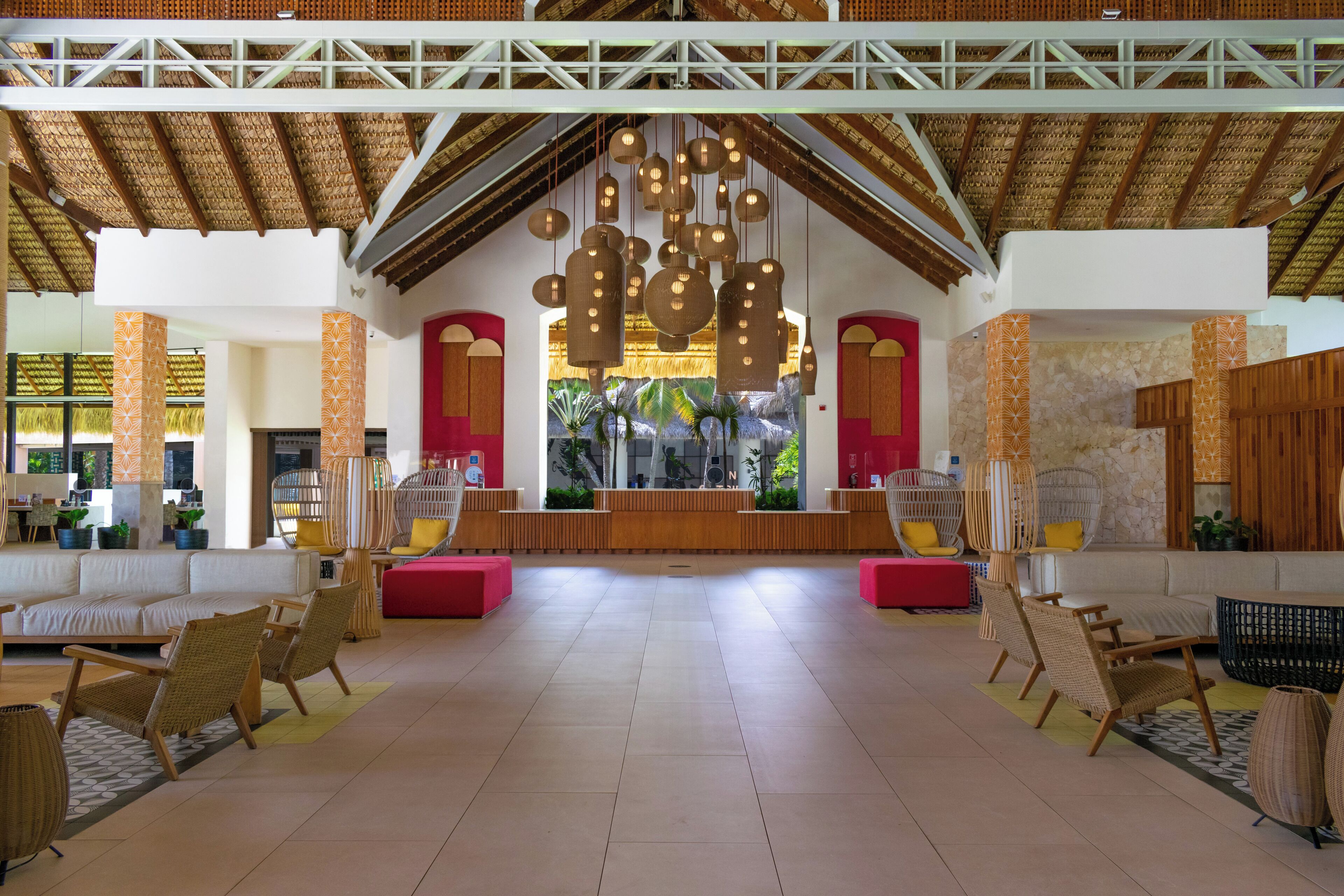 A spacious lobby with a high ceiling, wooden beams, and hanging spherical light fixtures. There are wicker chairs and sofas arranged around the space, with a reception desk in the center.