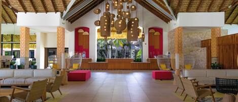 A spacious lobby with a high ceiling, wooden beams, and hanging spherical light fixtures. There are wicker chairs and sofas arranged around the space, with a reception desk in the center.