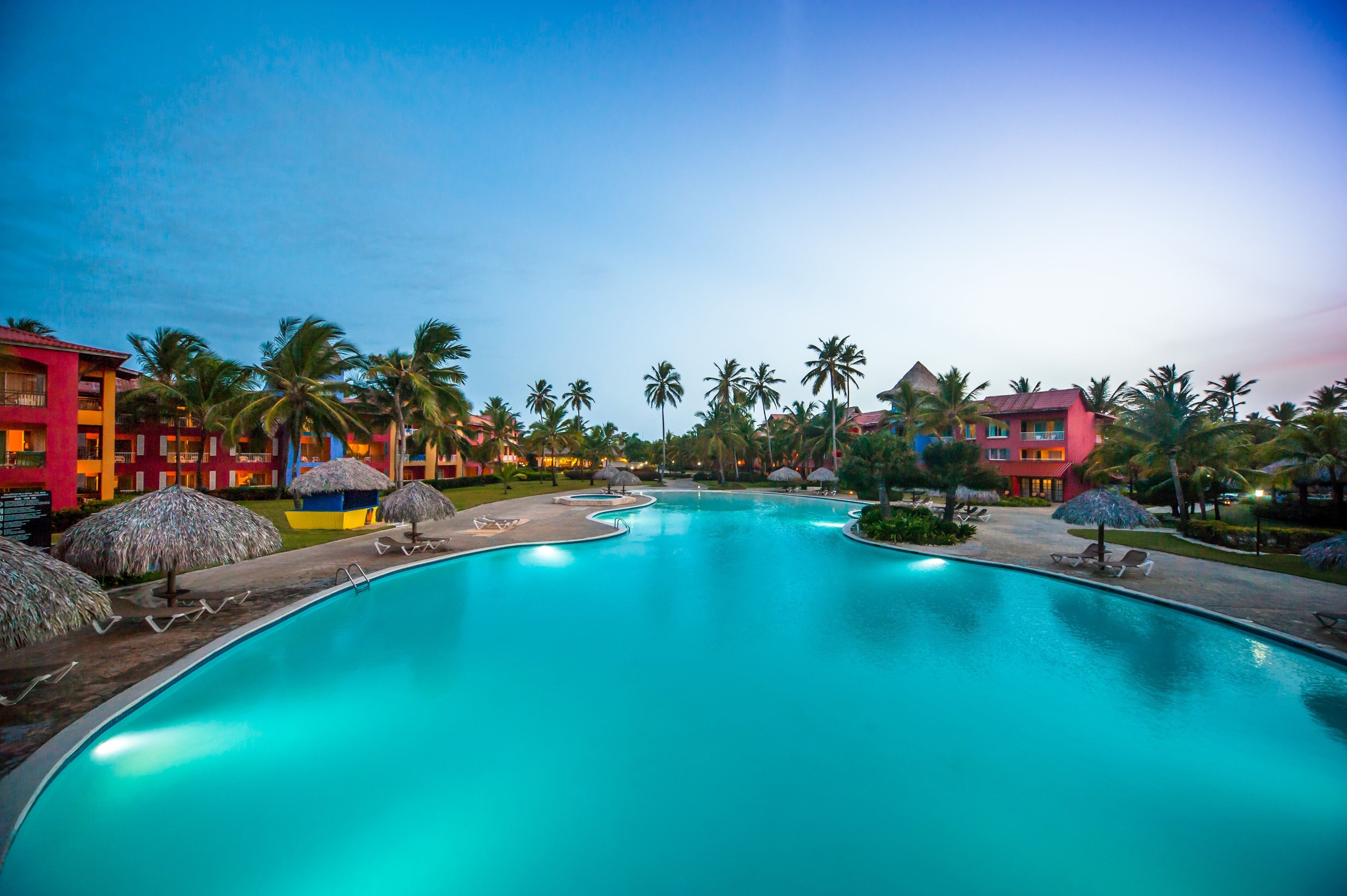 A swimming pool with lounge chairs and umbrellas, surrounded by palm trees and colorful buildings.