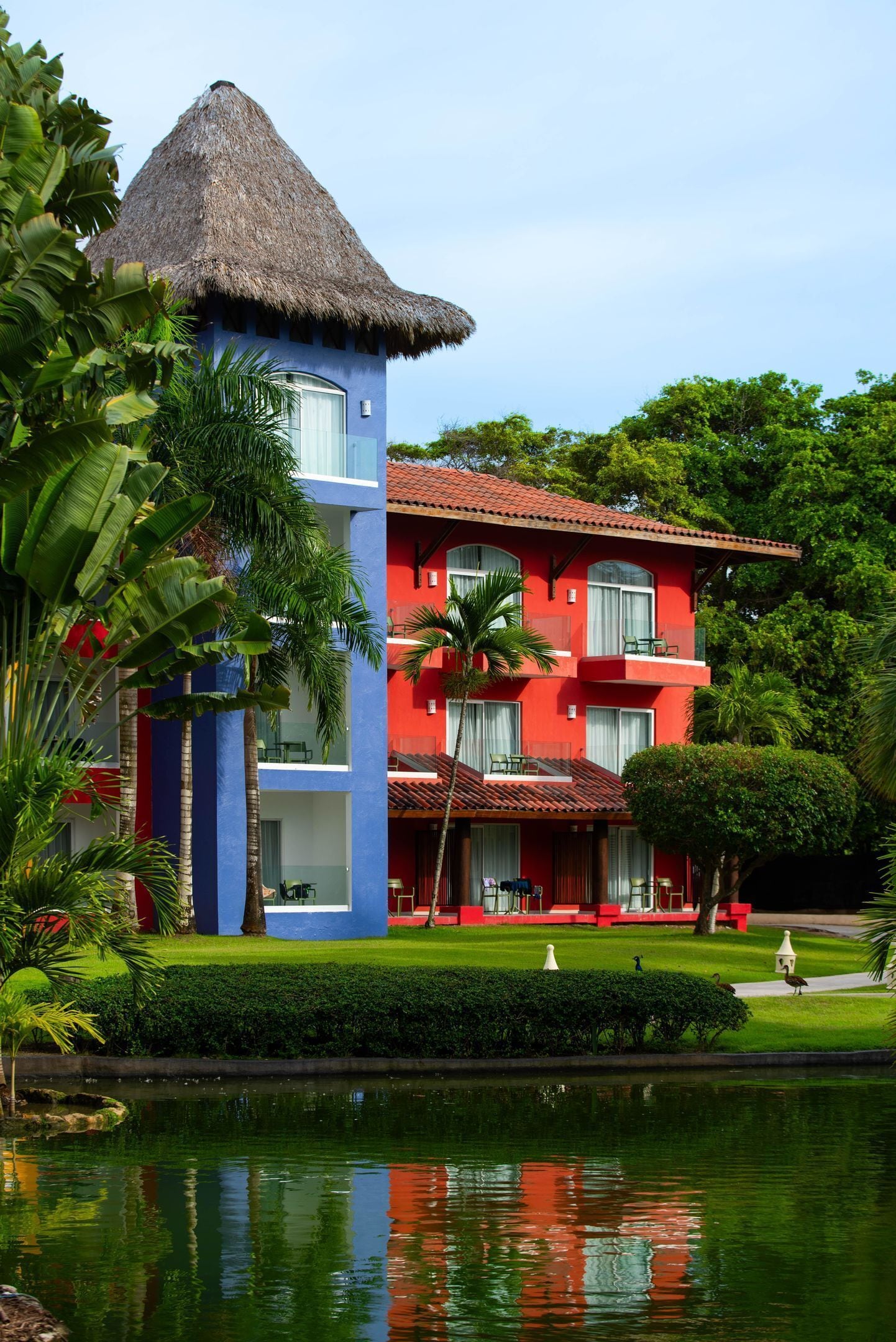 A multi-story building with a thatched roof, red and blue exterior, and a well-maintained garden with a pond.