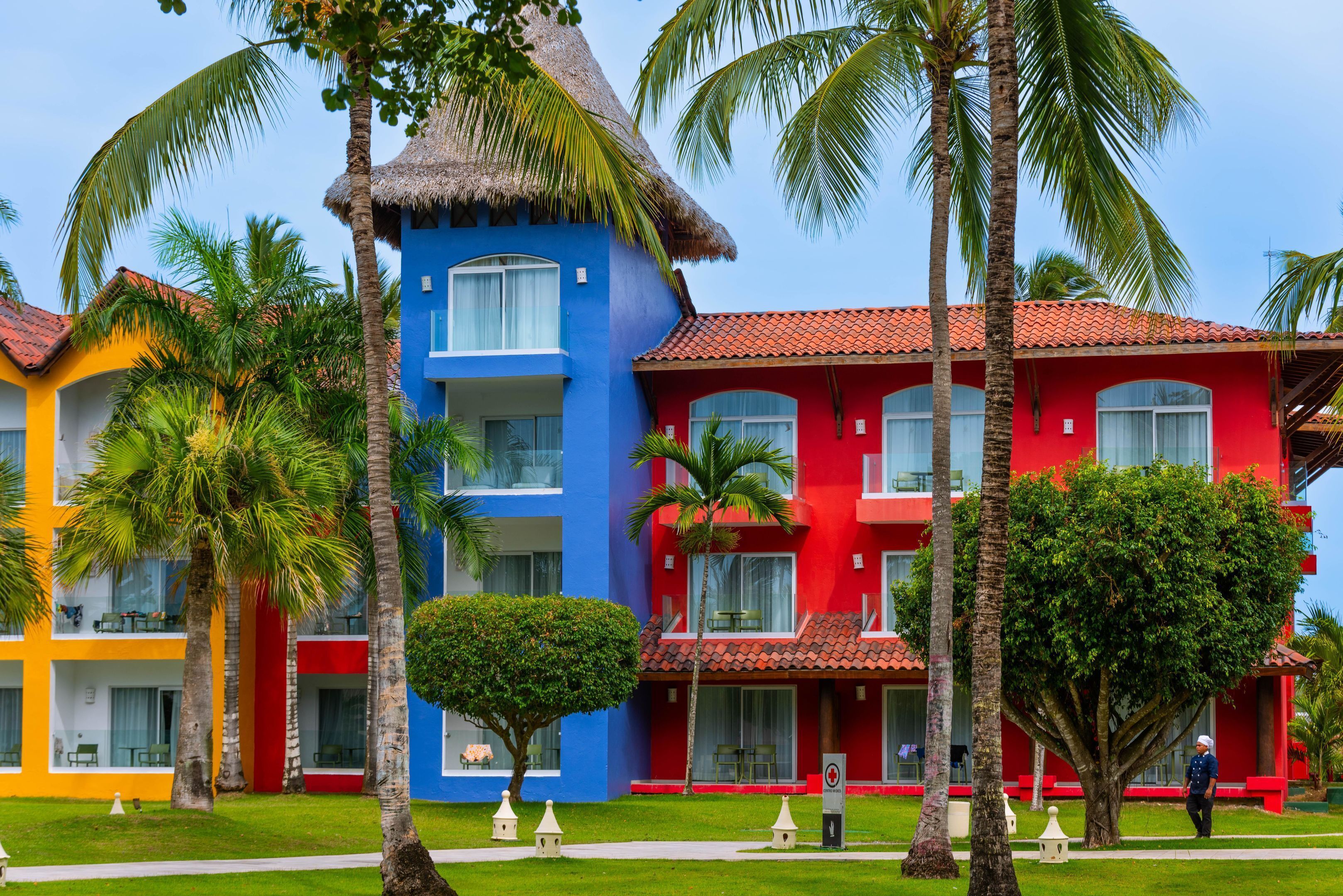 A colorful hotel with a blue building, red building, and a yellow building. There are palm trees and a well-maintained lawn.